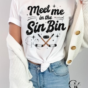 May include: White t-shirt with the text "Meet me in the Sin Bin" in black script. The design includes crossed hockey sticks, a puck, and star accents. The shirt is paired with blue jeans.