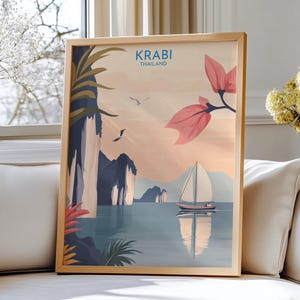 May include: A framed art print displaying a coastal scene with the text "KRABI THAILAND." The image shows a sailboat on tranquil waters, with mountains in the background and pink and green foliage. The frame is a light wood colour.
