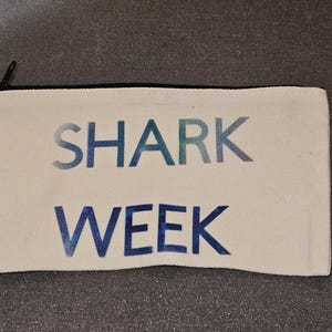 Op de afbeelding: Witte rechthoekige etui met de woorden "SHARK WEEK" gedrukt in blauwe en teal gradiënt letters. De etui heeft een zwarte rits en rand.