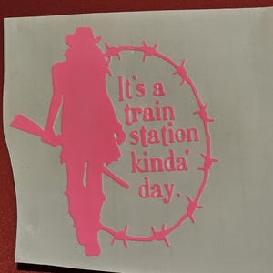 May include: A pink vinyl sticker featuring a silhouette of a person in a cowboy hat and holding a rifle, encircled by barbed wire. The text "It's a train station kinda' day" is inside the barbed wire circle.