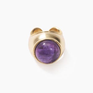 May include: A gold ring featuring a large, round, purple amethyst gemstone. The ring has a thick band and a rounded bezel setting. The gemstone is a deep shade of purple. The ring is set against a white background.