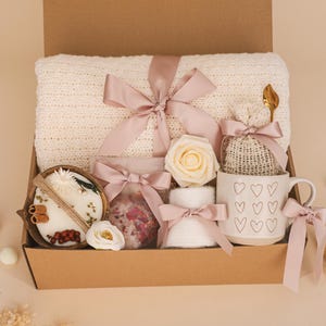 May include: A gift box filled with a cream-colored knitted blanket, a mug with heart designs, a candle, bath salts, a rose, and other items. The items are tied with pink ribbons.
