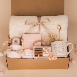 Op de afbeelding: Een cadeauverpakking gevuld met een crèmekleurige deken met een lint, een roze notitieboek, een kleine roze radio, een mok met hartjes, kaarsen en bloemen. De doos is bruin en staat op een beige ondergrond.