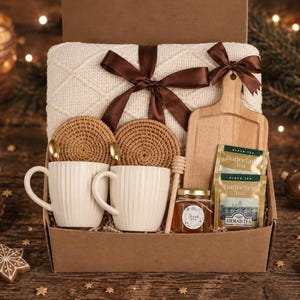 May include: A gift box containing a cream-coloured knit blanket tied with a brown ribbon, two ribbed white ceramic mugs, two round woven coasters, a small wooden cutting board, a jar of honey with a wooden dipper, and two tea bags labelled Darjeeling.