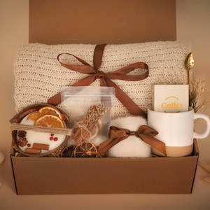 Könnte beinhalten: Eine braune Geschenkbox mit einer cremefarbenen Strickdecke, die mit einem braunen Band gebunden ist. Die Box enthält auch einen weiß-beigen Becher, eine Kerze mit getrockneten Orangenscheiben und einen kleinen, flauschigen Artikel mit einer Schleife.