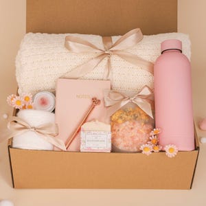 Pode incluir: Uma caixa de presente com itens de autocuidado. A caixa contém um cobertor creme, uma garrafa de água rosa, um caderno, uma caneta, uma vela, uma toalha pequena e sais de banho. Os itens são amarrados com fitas e decorados com flores.