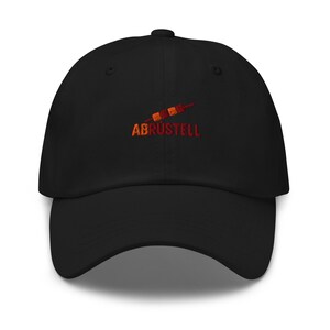 Può includere: Cappellino da baseball nero con visiera curva. Il cappellino presenta la scritta ricamata "ABRUSTELL" in arancione e rosso, insieme a un disegno stilizzato. Il cappellino è realizzato in tessuto morbido.