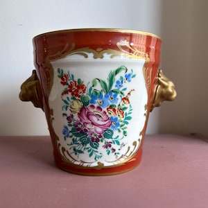 Peut inclure: Un vase décoratif en céramique avec un bord rouge et des accents dorés. Le vase présente un centre blanc avec un motif floral coloré. Des poignées dorées en forme de tête de lion sont de chaque côté.