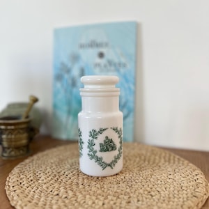 Peut inclure: Un pot d'apothicaire en céramique blanche avec un couvercle, décoré d'un emblème vert et d'une couronne de laurier. Le pot est cylindrique et repose sur un tapis tissé. Un livre est en arrière-plan.