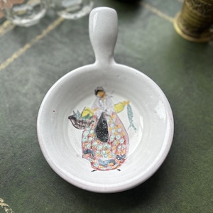 Könnte beinhalten: Kleine, weiße Keramikschale mit Griff. Die Schale zeigt eine handgemalte Illustration einer Person, die einen Korb und Fisch trägt. Die Person trägt ein farbenfrohes Kleid. Die Schale steht auf einer grünen Oberfläche.