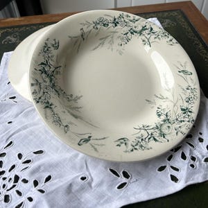 Peut inclure: Deux assiettes en céramique blanc cassé avec un motif floral et d'oiseaux en nuances de vert et de gris. Les assiettes sont empilées sur un tissu blanc à œillets, créant une esthétique vintage. Les assiettes ont une forme peu profonde.