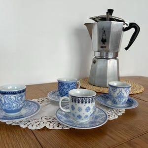 Peut inclure: Quatre tasses et soucoupes à expresso bleues et blanches avec des motifs floraux ornent une nappe en dentelle blanche. Une cafetière italienne en argent est posée sur un tapis tressé.