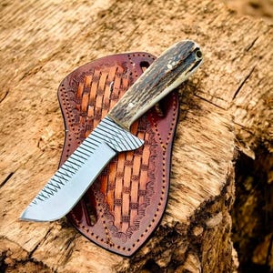 May include: A fixed-blade knife with a textured metal blade featuring a cross-hatched pattern near the bolster and a natural antler handle. The knife rests on a brown leather sheath with a decorative basket-weave pattern and stitched edges.
