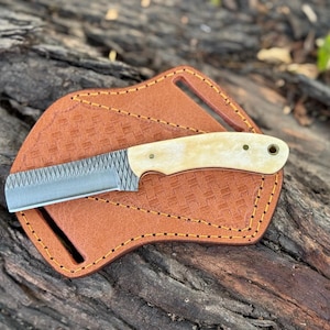 May include: A small, handcrafted knife with a textured blade and a cream-colored handle rests on a brown leather sheath. The sheath is stitched with yellow thread and has a basketweave pattern. The knife and sheath are displayed on a weathered wooden surface.
