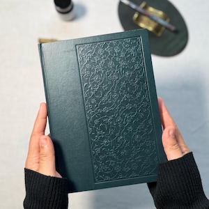 May include: A dark teal journal with an embossed floral design on the cover. The journal is held open, showcasing the intricate pattern. The cover has a smooth texture, and the edges are clean and well-defined.