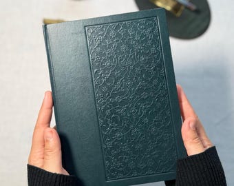 May include: A dark teal journal with an embossed floral design on the cover. The journal is held open, showcasing the intricate pattern. The cover has a smooth texture, and the edges are clean and well-defined.