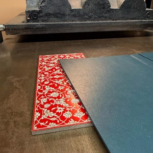 May include: A red and white patterned decorative piece with a floral design, next to a blue-gray piece of material. The items are on a wooden surface, with a large black printing press in the background.