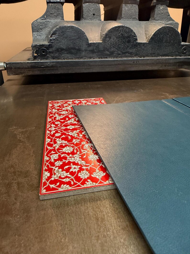 May include: A red and white patterned decorative piece with a floral design, next to a blue-gray piece of material. The items are on a wooden surface, with a large black printing press in the background.