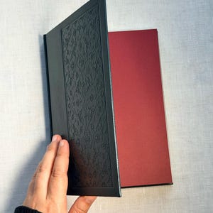May include: A black book with an embossed floral design on the cover is held open, revealing a red interior page. The book's cover is textured, and the pages are a contrasting color.