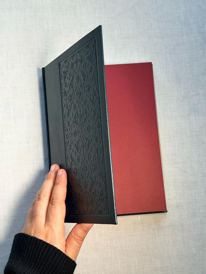 May include: A black book with an embossed floral design on the cover is held open, revealing a red interior page. The book's cover is textured, and the pages are a contrasting color.
