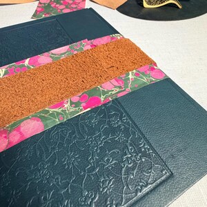 May include: A close-up of a dark teal leather journal with embossed floral patterns. The journal is adorned with strips of marbled paper in pink and green, and a textured brown leather strip. Additional leather pieces and a pen are visible.