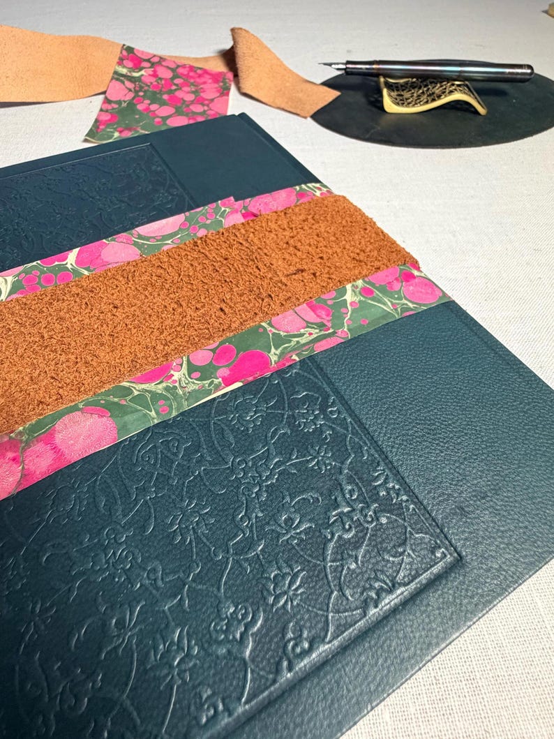 May include: A close-up of a dark teal leather journal with embossed floral patterns. The journal is adorned with strips of marbled paper in pink and green, and a textured brown leather strip. Additional leather pieces and a pen are visible.