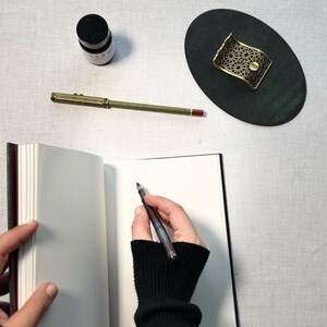May include: A vintage-style pen with a gold-colored barrel and a dark ink bottle sit on a white surface. A hand wearing a black sleeve is holding the pen over an open notebook. A decorative brass pen holder is also present.