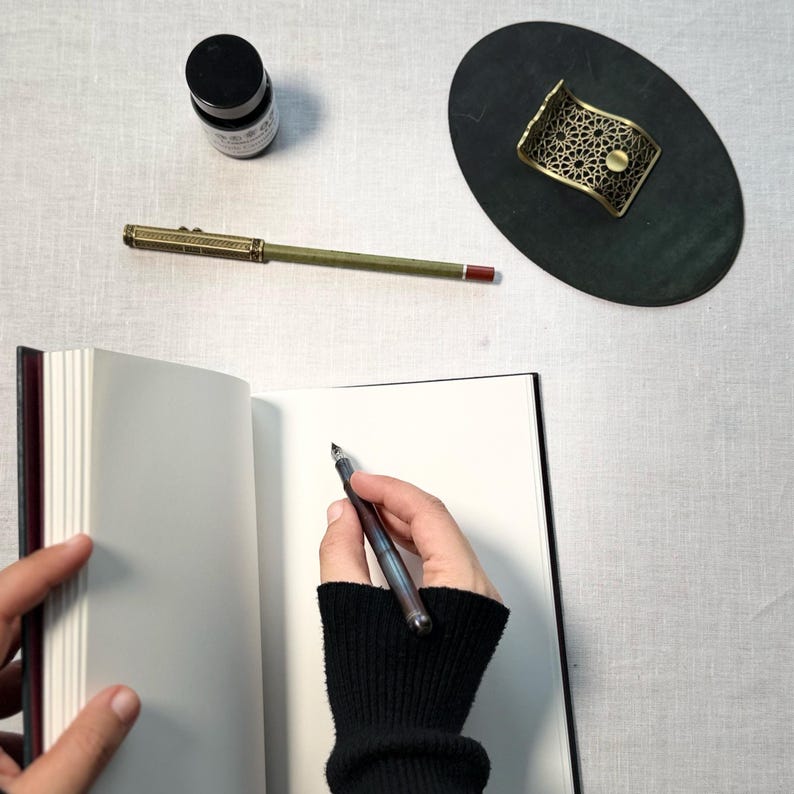 May include: A vintage-style pen with a gold-colored barrel and a dark ink bottle sit on a white surface. A hand wearing a black sleeve is holding the pen over an open notebook. A decorative brass pen holder is also present.