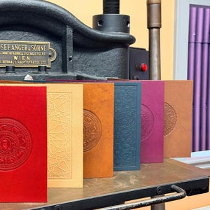 May include: A collection of embossed journals in various colors, including red, yellow, orange, blue, purple, and brown. Each journal features a unique circular design. The journals are displayed on a metal surface with a vintage printing press in the background.