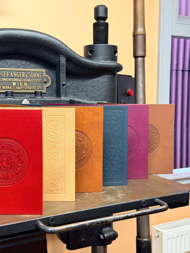 May include: A collection of embossed journals in various colors, including red, yellow, orange, blue, purple, and brown. Each journal features a unique circular design. The journals are displayed on a metal surface with a vintage printing press in the background.