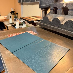 May include: A teal-colored, open book or folio with an embossed floral design on the right panel, resting on a wooden surface. The image is taken in a workshop setting, with a printing press visible in the background.