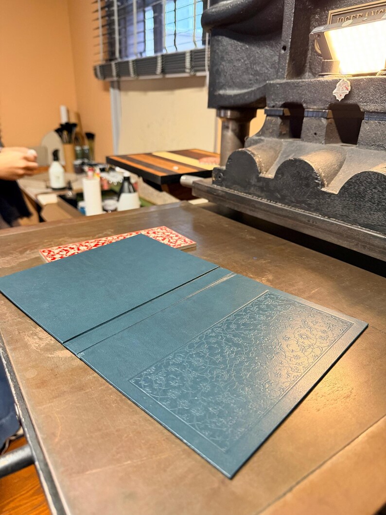May include: A teal-colored, open book or folio with an embossed floral design on the right panel, resting on a wooden surface. The image is taken in a workshop setting, with a printing press visible in the background.