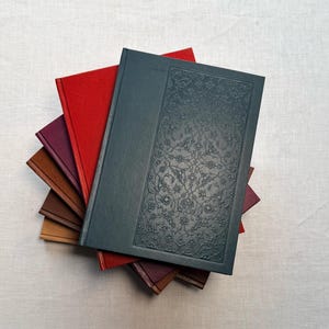 May include: A stack of hardcover books with various colored covers. The top book is dark teal with an embossed floral design. Other books have red, burgundy, and brown covers. The books are arranged on a light-colored fabric surface.