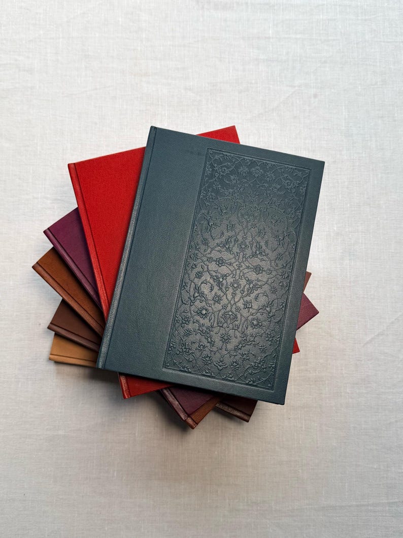 May include: A stack of hardcover books with various colored covers. The top book is dark teal with an embossed floral design. Other books have red, burgundy, and brown covers. The books are arranged on a light-colored fabric surface.