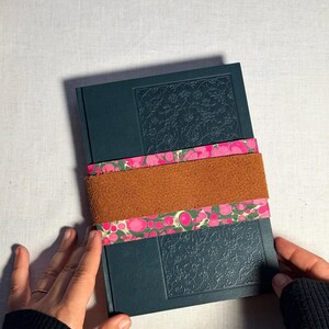 May include: A dark teal journal with embossed floral detailing, secured by a brown suede band and a pink and green patterned paper band. The journal is held by hands wearing a black sleeve.