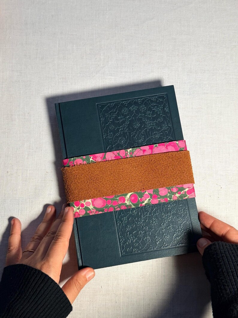 May include: A dark teal journal with embossed floral detailing, secured by a brown suede band and a pink and green patterned paper band. The journal is held by hands wearing a black sleeve.