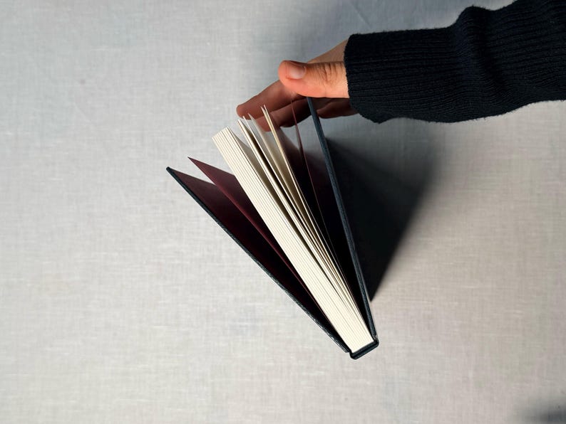 May include: A hardcover book with a dark blue cover and white pages is held open, revealing a burgundy interior. The book's pages are fanned out, and a hand is visible holding the book. The background is a light gray surface.