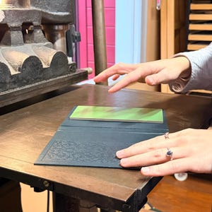 May include: A printing press with a dark gray metal frame and a wooden surface. A dark blue book with an embossed floral design is open on the press. A green and brown rectangular object rests inside the book. The scene is illuminated by a bright overhead light.