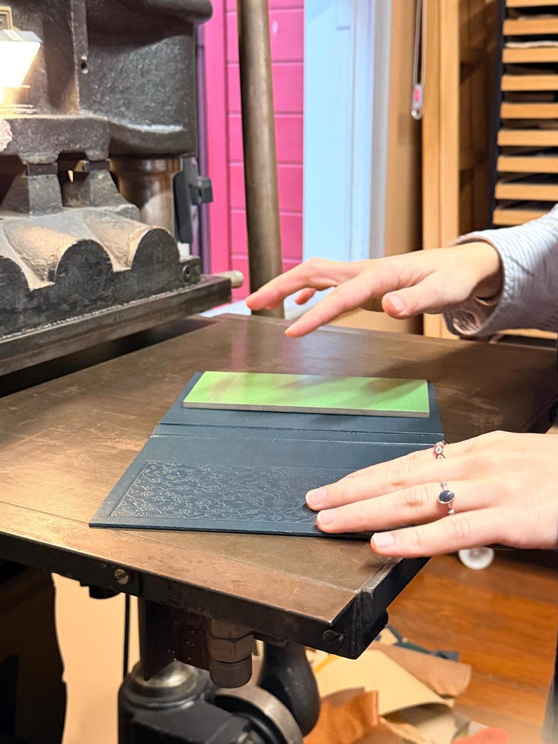 May include: A printing press with a dark gray metal frame and a wooden surface. A dark blue book with an embossed floral design is open on the press. A green and brown rectangular object rests inside the book. The scene is illuminated by a bright overhead light.