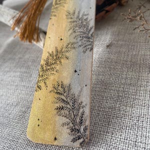 Può includere: Un segnalibro in legno con una sfumatura da oro a argento, con un design dettagliato di felce. Il segnalibro ha un nappa marrone attaccata. Un accessorio di lettura ispirato alla natura.