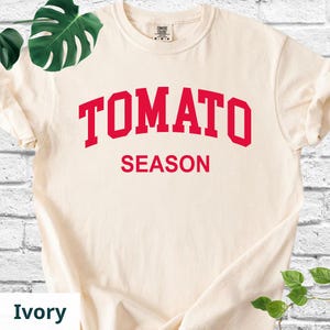 K&ouml;nnte beinhalten: Elfenbeinfarbenes T-Shirt mit dem roten Aufdruck "TOMATO SEASON" in fetter Schrift. Das T-Shirt hat einen Rundhalsausschnitt und kurze &Auml;rmel. Der Text ist in einer Schriftart im College-Stil gehalten. Das T-Shirt ist ein l&auml;ssiger Stil.