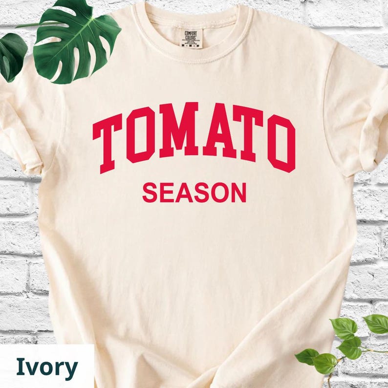 K&ouml;nnte beinhalten: Elfenbeinfarbenes T-Shirt mit dem roten Aufdruck "TOMATO SEASON" in fetter Schrift. Das T-Shirt hat einen Rundhalsausschnitt und kurze &Auml;rmel. Der Text ist in einer Schriftart im College-Stil gehalten. Das T-Shirt ist ein l&auml;ssiger Stil.