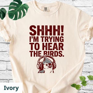 May include: An ivory short-sleeve t-shirt featuring the dark red text SHHH! I'M TRYING TO HEAR THE BIRDS. Below the text is a graphic of a bird wearing large headphones. The shirt has a crew neckline and a standard fit, made of cotton fabric.