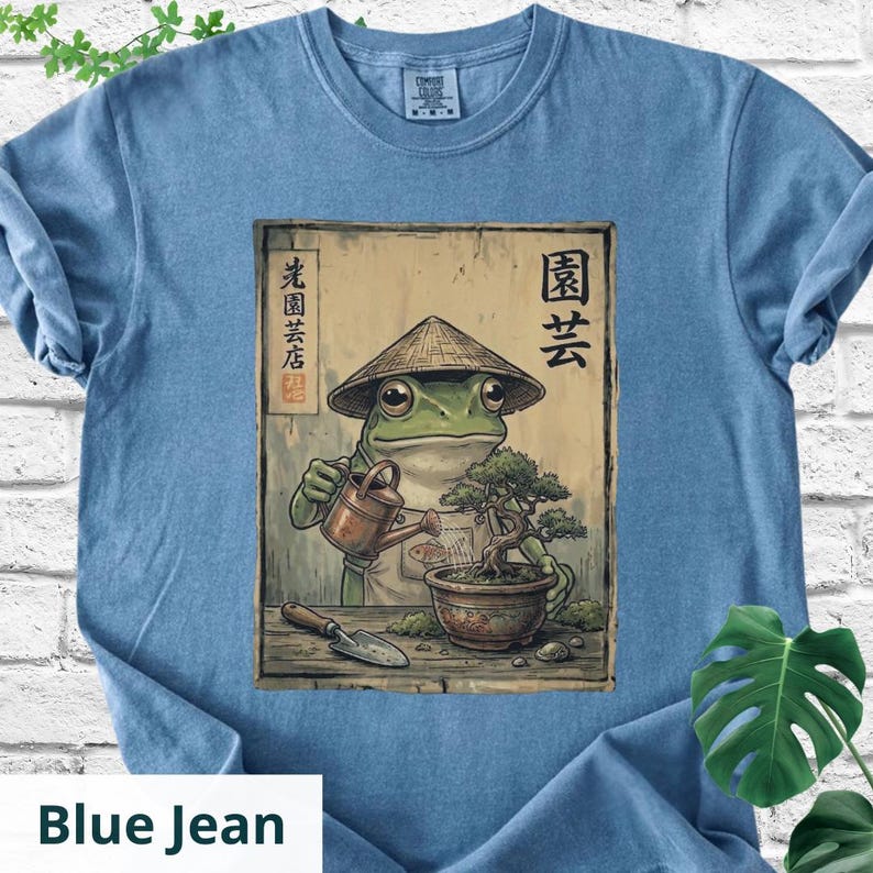 May include: Blue jean colored t-shirt featuring a frog in a conical hat watering a bonsai tree. The frog wears an apron and holds a watering can. Japanese text and a small shovel are also visible.