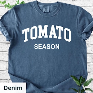 K&ouml;nnte beinhalten: Denimblaues T-Shirt mit dem wei&szlig;en, fettgedruckten Schriftzug "TOMATO SEASON". Das Shirt hat einen Rundhalsausschnitt und kurze &Auml;rmel. Der Stoff wirkt weich und leicht verwaschen, was ihm einen Vintage-Look verleiht. Ein l&auml;ssiges Kleidungsst&uuml;ck.