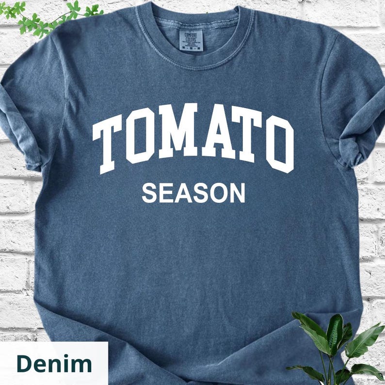K&ouml;nnte beinhalten: Denimblaues T-Shirt mit dem wei&szlig;en, fettgedruckten Schriftzug "TOMATO SEASON". Das Shirt hat einen Rundhalsausschnitt und kurze &Auml;rmel. Der Stoff wirkt weich und leicht verwaschen, was ihm einen Vintage-Look verleiht. Ein l&auml;ssiges Kleidungsst&uuml;ck.
