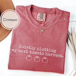 May include: Crimson-colored t-shirt with the text "Quietly plotting my next tomato harvest" and three tomato illustrations. A silver necklace with a pendant rests on the shirt. The shirt is a Comfort Colors brand.