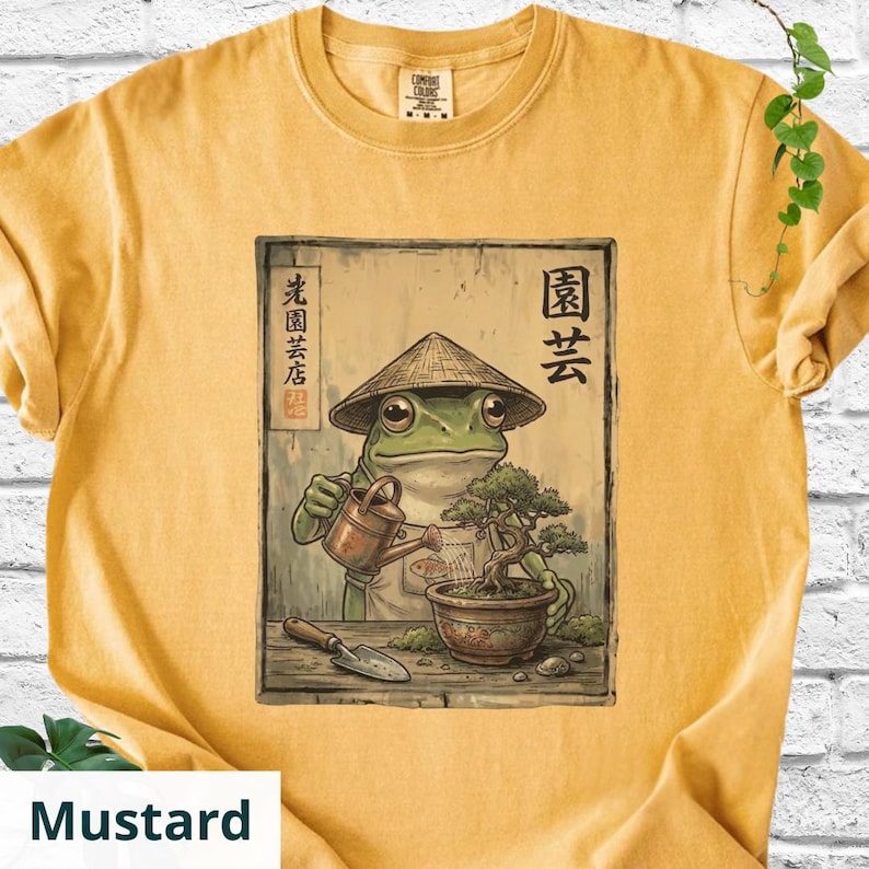 May include: Mustard-colored t-shirt featuring a framed illustration of a frog wearing a conical hat, watering a bonsai tree. The frog is holding a watering can. Japanese text is visible. The shirt has a crew neck and short sleeves.