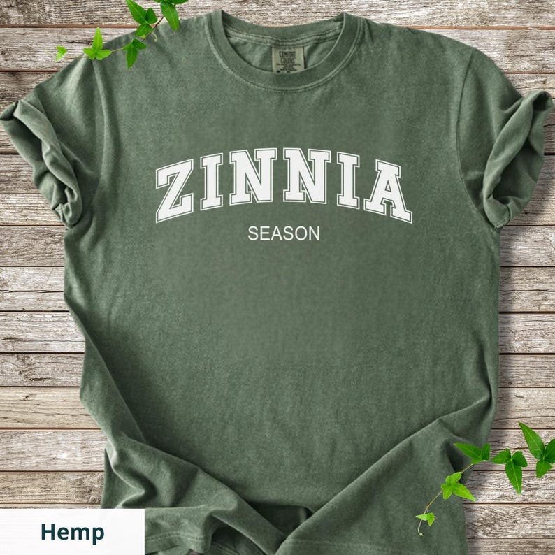 May include: A green t-shirt featuring the white text "ZINNIA" above "SEASON". The shirt has rolled-up sleeves and is made of a soft material. The word "Hemp" is at the bottom of the image.
