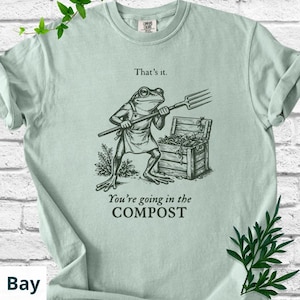 May include: A light green t-shirt featuring a frog illustration holding a pitchfork, standing near a wooden compost bin. The text above the frog reads "That's it." and below it, "You're going in the COMPOST."